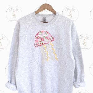 May include: A gray sweatshirt with a floral patterned jellyfish appliqué on the front. The jellyfish is pink, white, and yellow.