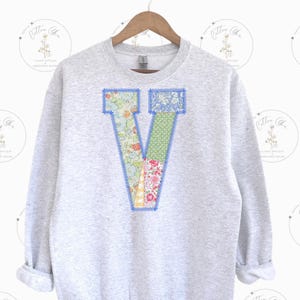 May include: A gray sweatshirt with a large patchwork letter V on the front. The letter is made of different floral fabrics and is outlined in blue.