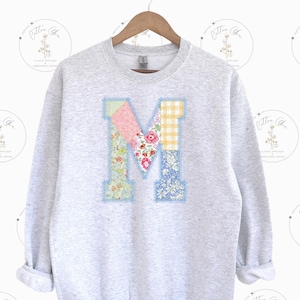 May include: A gray sweatshirt with a large patchwork letter "M" on the front. The letter is made of different floral and checkered fabrics in shades of pink, blue, yellow, and green.