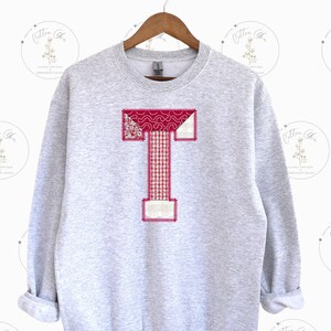 May include: A gray sweatshirt with a large red, white, and pink patchwork letter T on the front.
