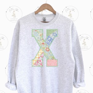 May include: A gray sweatshirt with a large patchwork letter X on the front. The letter is made of different floral fabrics in shades of pink, blue, green, and yellow.