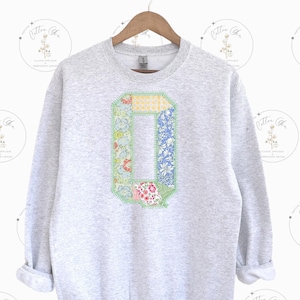 May include: A gray sweatshirt with a large patchwork letter Q on the front. The letter is made of different floral fabrics and is outlined in green.