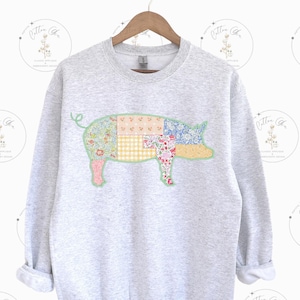 Peut inclure: Un sweat-shirt gris clair avec un motif de cochon en patchwork. Le cochon est composé de divers tissus à motifs floraux et autres, dans les tons de rose, bleu, jaune et vert. Le sweat-shirt a un col rond et des manches longues.