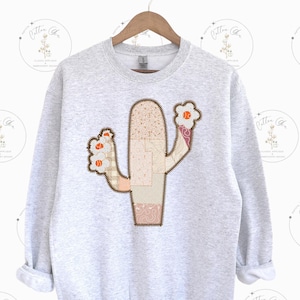 May include: A gray sweatshirt with a patchwork cactus design in shades of pink, beige, and brown. The cactus has small orange flowers.