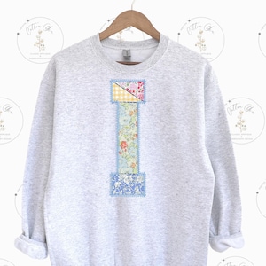 May include: A gray sweatshirt with a patchwork letter I on the front. The letter is made of different floral and checkered fabrics in shades of blue, yellow, pink, and white.