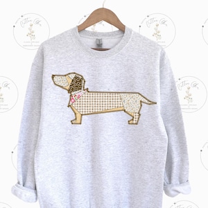 Puede incluir: Una sudadera gris claro con un diseño de perro salchicha en patchwork. El perro está compuesto por varias telas estampadas en tonos marrones, beige y rosa. La sudadera tiene cuello redondo y mangas largas.