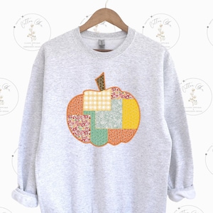 Peut inclure: Un sweat-shirt gris clair orné d'un motif de citrouille en patchwork. La citrouille est composée de divers motifs de tissus orange, jaune, vert et à imprimé floral. Le sweat-shirt a un col rond et des manches longues.