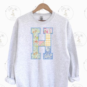 Könnte beinhalten: Ein grauer Pullover mit einem großen Patchwork-Buchstaben H auf der Vorderseite. Der Buchstabe besteht aus verschiedenen Blumenstoffen in den Farben Rosa, Blau, Gelb und Grün.