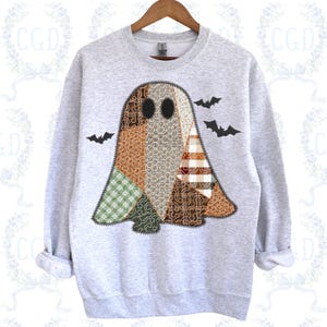 Puede incluir: Una sudadera gris claro con un diseño de fantasma de patchwork, ojos negros y un detalle de cremallera. El fantasma está compuesto por varios patrones de tela en tonos marrones, verdes y blancos. Tres siluetas de murciélagos negros están dispersas.