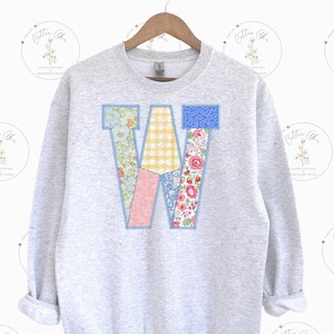 May include: A light gray sweatshirt with a large, patchwork letter "W" on the front. The letter is composed of various floral and patterned fabrics in shades of blue, yellow, pink, and red, stitched together with blue thread.