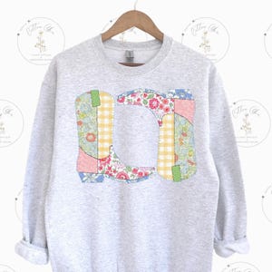 May include: A light gray sweatshirt featuring a colorful patchwork design of cowboy boots. The boots are composed of floral, gingham, and patterned fabrics in shades of pink, blue, yellow, and green. The sweatshirt has a crew neck and long sleeves.