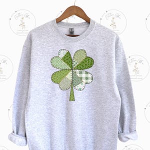 May include: A grey sweatshirt featuring a green four-leaf clover design. The clover is made up of different green patterned fabric pieces.
