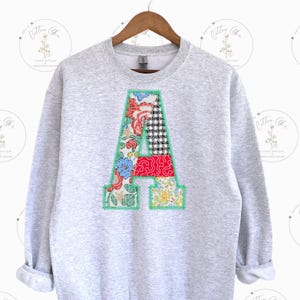 May include: A gray sweatshirt with a large patchwork letter A on the front. The letter is made of different floral fabrics in shades of red, blue, green, and yellow.