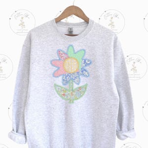 May include: A gray sweatshirt with a colorful patchwork flower design. The flower is made of different fabrics and has a green stem and leaves.