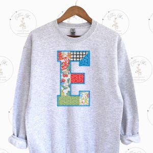 May include: A light gray sweatshirt with a large, colorful letter "E" appliqued on the front. The letter is made of various patterned fabrics, including floral, checkered, and abstract designs, outlined with blue stitching.