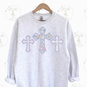 Könnte beinhalten: Ein grauer Pullover mit drei floralen Kreuzdesigns in Blautönen, Rosa und Grün.