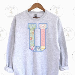 May include: A gray sweatshirt with a large patchwork letter U in blue, pink, and green floral fabric. The letter is outlined in blue stitching.