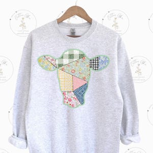 May include: A gray sweatshirt with a patchwork cow design in shades of green, pink, blue, and yellow. The cow is made up of different fabric scraps sewn together.