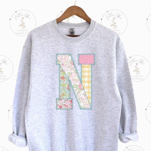 May include: A gray sweatshirt with a large patchwork letter N in pink, yellow, and green floral patterns. The letter is outlined in blue thread.