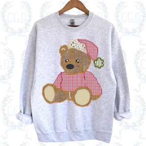 May include: A light gray sweatshirt featuring a teddy bear design. The bear wears a red and white gingham shirt and a Santa hat. The bear's design is stitched with brown, tan, and black thread. The sweatshirt is on a wooden hanger.
