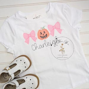 Pode incluir: Uma camiseta branca com laços rosa e uma abóbora com um rosto de abóbora esculpido bordado nela. O texto "Charleigh" está bordado abaixo da abóbora. O texto "Classic Applique Embroidery Design" está bordado em um círculo no canto inferior direito da camiseta.