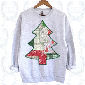 May include: A light gray sweatshirt with a Christmas tree design. The tree is made of green, red, and white patchwork fabric. The tree is stitched onto the sweatshirt with a zig-zag stitch. The sweatshirt is hanging on a wooden hanger.