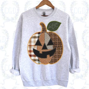 May include: A light gray sweatshirt featuring a patchwork pumpkin design. The pumpkin has a black carved face, a green leaf, and is made of various patterned fabrics in shades of brown, orange, and cream. The sweatshirt is hanging on a wooden hanger.