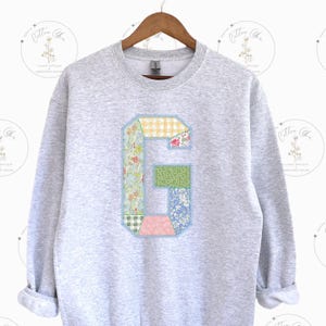 May include: A gray sweatshirt with a large patchwork letter G on the front. The letter is made of different floral and checkered fabrics in shades of pink, blue, green, and yellow.