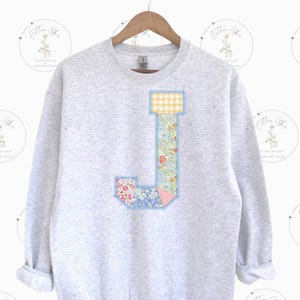 May include: A gray sweatshirt with a large patchwork letter J on the front. The letter is made of different floral and gingham fabrics.