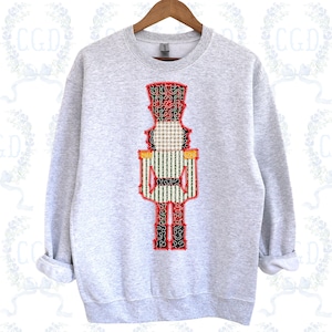 以下が含まれることがあります： カラフルなくるみ割り人形のデザインが施されたライトグレーのスウェットシャツ。くるみ割り人形は、赤、緑、黒、金のアクセントで詳細に描かれています。デザインはスウェットシャツの前面中央に配置され、長袖とクルーネックが付いています。