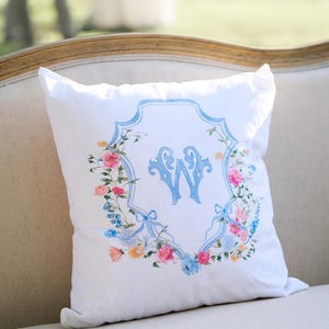 May include: White square pillow with a floral wreath design and a light blue monogram letter "W". The wreath features pink, blue, and yellow flowers with light blue ribbons. The pillow is on a light-colored sofa.