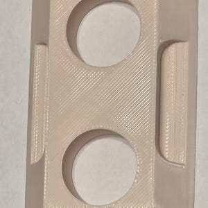 May include: A light beige, rectangular object with two circular openings and a raised, rectangular section on the side. The object has a textured surface with diagonal lines and rounded corners. It appears to be a 3D-printed item.