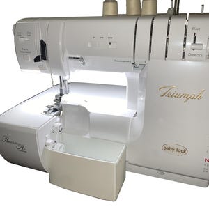 May include: A white Baby Lock Triumph serger sewing machine. The machine has a built-in light, a thread catcher, and various control dials. The Baby Lock logo is visible, along with the Triumph name in gold script.