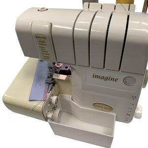 May include: A cream-colored Baby Lock Imagine serger sewing machine. The machine features the word "imagine" and the Baby Lock logo. A piece of blue fabric with a pink and purple stitch is visible. The machine has multiple dials and a white waste bin.