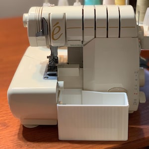 May include: Off-white serger sewing machine with a rectangular waste bin attached. The machine has the brand name "intimate" and the letter "é" in gold. The machine is on a wooden surface.