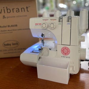 May include: A white Baby Lock BL4600D serger machine with a vibrant red and white logo. The machine has multiple dials, thread spools, and a built-in light. A cardboard box with the word "vibrant" is in the background.