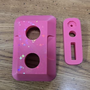 May include: Two-piece set of pink 3D-printed objects. The larger piece has two circular cutouts and holographic star accents. The smaller piece is rectangular with a hole and a slot. The objects are on a wooden surface.