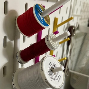 May include: Close-up of a white pegboard with three spools of thread. The spools are red, dark red, and white, with white plastic spool holders. The thread spools are labeled with brand names and thread weights.
