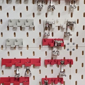 May include: A collection of sewing machine presser feet and holders, displayed on a white pegboard. The holders are in red, white, and gray, with the presser feet in silver. The pegboard has evenly spaced holes.