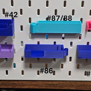 May include: Collection of colorful 3D-printed storage containers in pink, blue, purple, and teal. Each container has a unique shape and design, with numbers printed on them. The containers are mounted on a white pegboard.