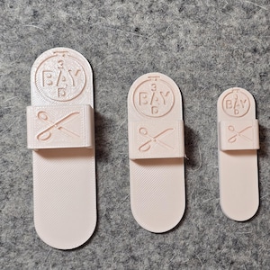 May include: Three light pink, 3D-printed bookmarks. Each bookmark has a rectangular section with scissors and the text "BAY" above it. The bookmarks are different sizes, with the largest on the left and the smallest on the right.