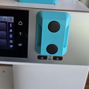 May include: Close-up of a sewing machine with a light blue control panel and black buttons. The machine has a digital display and the brand name "BERNINA" is visible in red. The machine is white and silver.