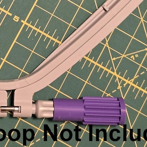 May include: A gray embroidery hoop with a purple adjustment tool, set against a green cutting mat with yellow grid lines. The text "Hoop Not Included" is visible.