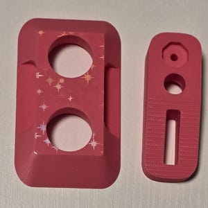 May include: Two bright pink, 3D-printed components. One is rectangular with two circular cutouts and star-shaped holographic accents. The other is a smaller, elongated piece with a hole and a rectangular cutout.