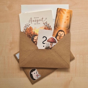 Könnte beinhalten: Ein brauner Kraftpapierumschlag mit einer Grußkarte mit dem Text "Kleine Auszeit für dich" und einer Igel-Illustration, einem Lesezeichen mit Waldmotiv und Igel sowie einer kleinen Karte mit einem Fragezeichen und einem Fliegenpilz.
