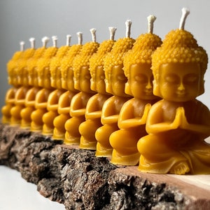 Könnte beinhalten: Eine Reihe kleiner, gelber Bienenwachskerzen in Form von meditierenden Buddha-Figuren. Die Kerzen sind auf einem Stück Baumrinde angeordnet.