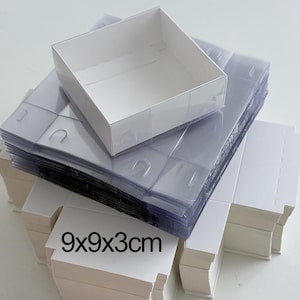 Könnte beinhalten: Ein Stapel weißer Kartons und transparenter Kunststoffhüllen. Auf dem Stapel befindet sich eine weiße Box. Der Text "9x9x3cm" ist auf die Vorderseite des weißen Kartons gedruckt.