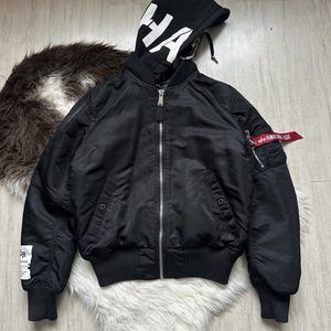 May include: Black bomber jacket with a black hood featuring the word "HA" in white. The jacket has a silver zipper, ribbed cuffs, and a red tag on the sleeve. The jacket is displayed on a white fluffy surface.