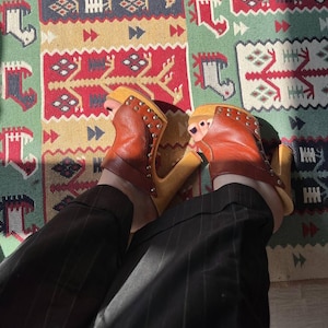 Pode incluir: Tamancos de couro castanho com design peep-toe e detalhes com tachas. Os tamancos têm um salto plataforma de madeira. A pessoa está usando calças pretas com riscas de giz. O fundo é um tapete estampado colorido.