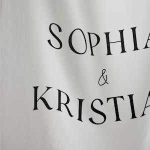 May include: A white fabric banner featuring the black printed text SOPHIA & KRISTIAN. The material has a visible woven texture and soft folds, with the names arranged in two lines separated by an ampersand in a classic serif typeface.